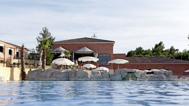 A pool area with a building in the background, featuring outdoor seating and umbrellas.