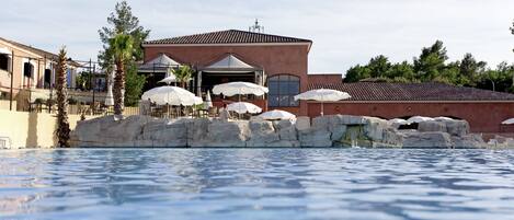 Zona de piscina con un edificio al fondo, con mobiliario exterior y sombrillas.