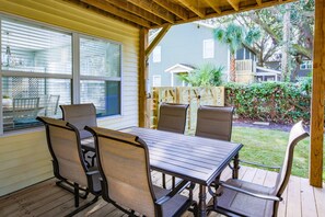 Une terrasse couverte, avec une table en bois et quatre chaises, donnant sur un jardin avec une clôture en bois et de la verdure.