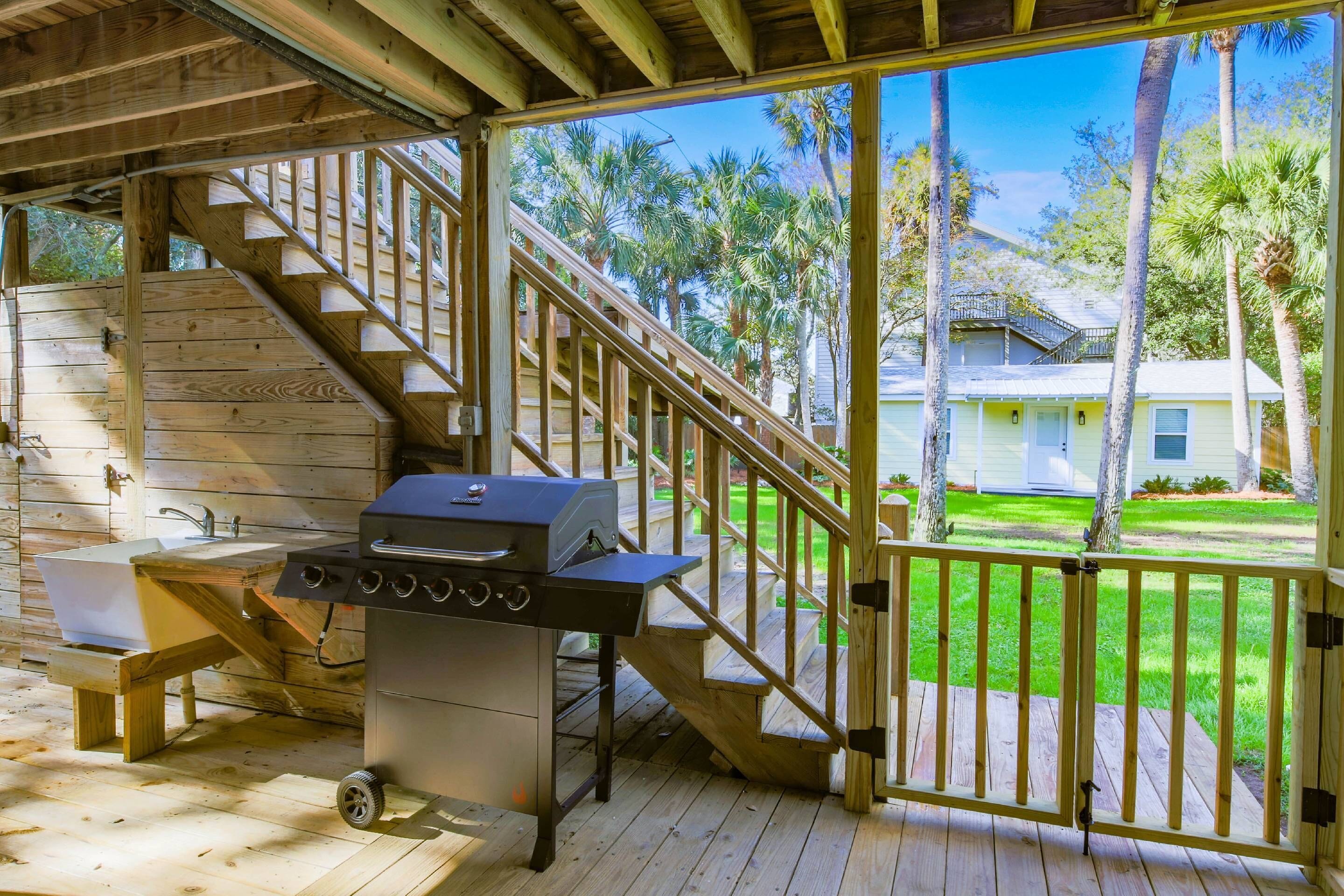A covered outdoor area with a wooden deck, a grill, a sink, and a staircase leading to a house.