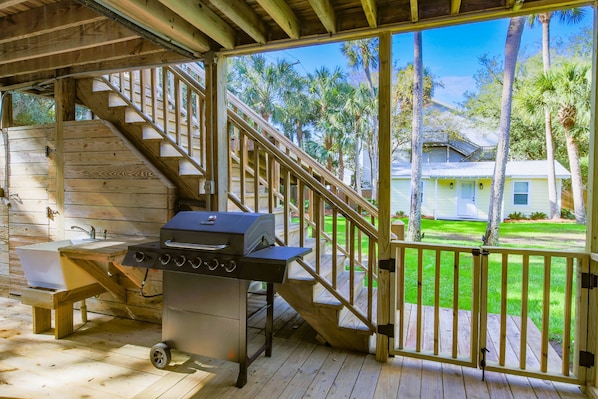 Un espacio exterior techado con terraza de madera, una parrilla, un lavamanos y una escalera que lleva a una casa.