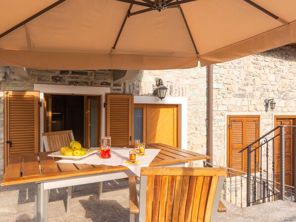 An outdoor dining area with a table set for two, a wooden bench, and a large umbrella providing shade.