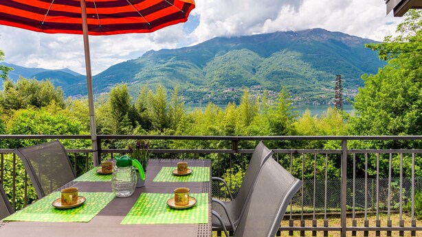 Eine Terrasse mit Tisch für zwei Personen, einem gestreiften Sonnenschirm und Blick auf Berge und einen See.