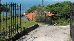 Ein Kiesweg führt zu einem Haus mit roten Ziegeln, umgeben von viel Grün und mit Blick auf einen See mit Bergen im Hintergrund.