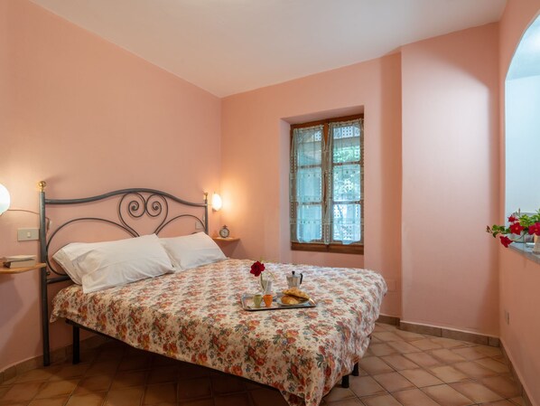 Une chambre à coucher avec une couvre-lit à motifs floraux, une table de chevet avec une lampe, une horloge et une fenêtre ornée de rideaux en dentelle.
