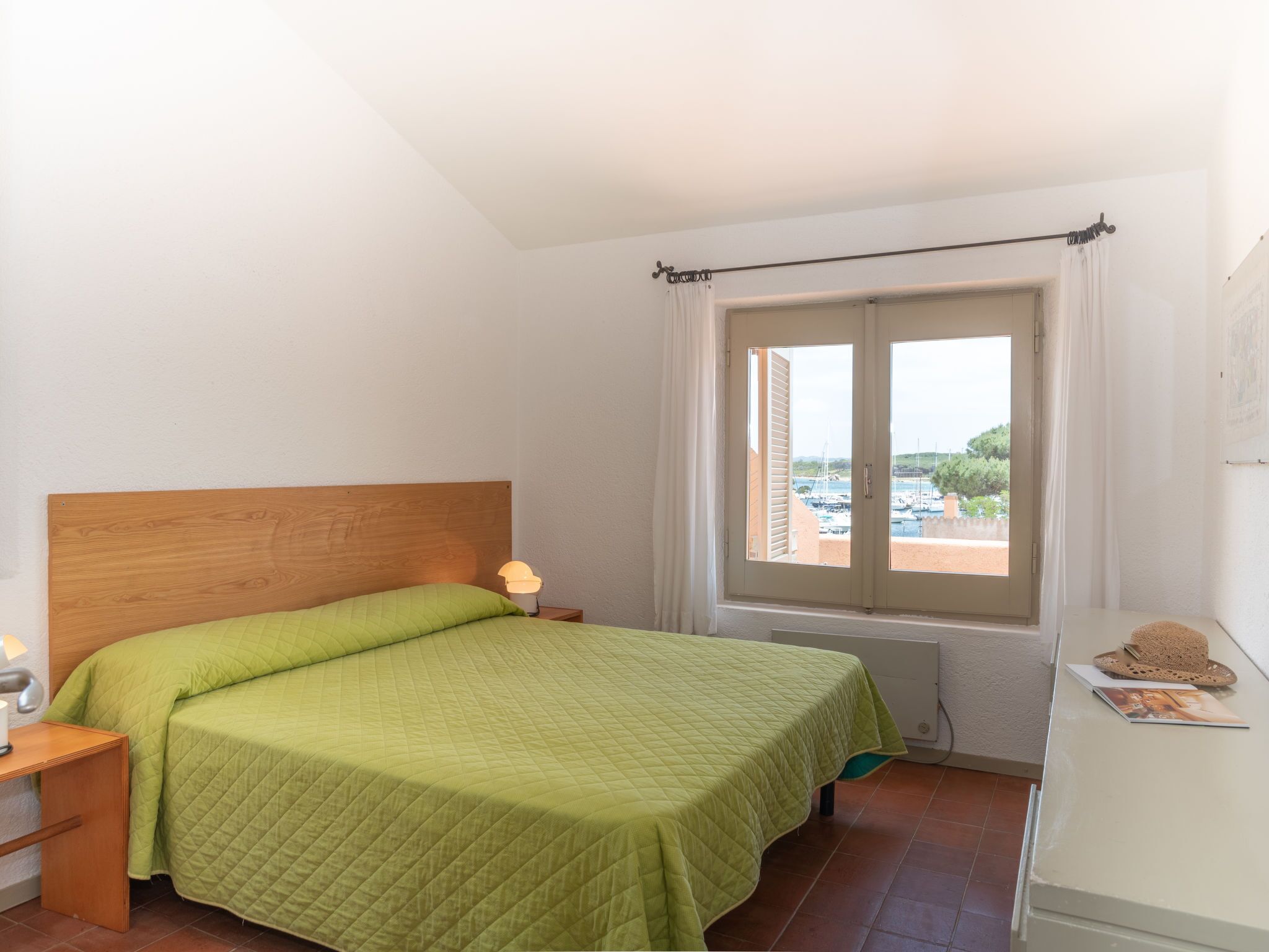Ein Schlafzimmer mit einem hölzernen Kopfteil, einer gesteppten grünen Bettdecke, einer Nachttischlampe, einem Fenster mit Blick auf Boote und Grünflächen sowie einem kleinen Tisch mit Hut und Bilderrahmen.