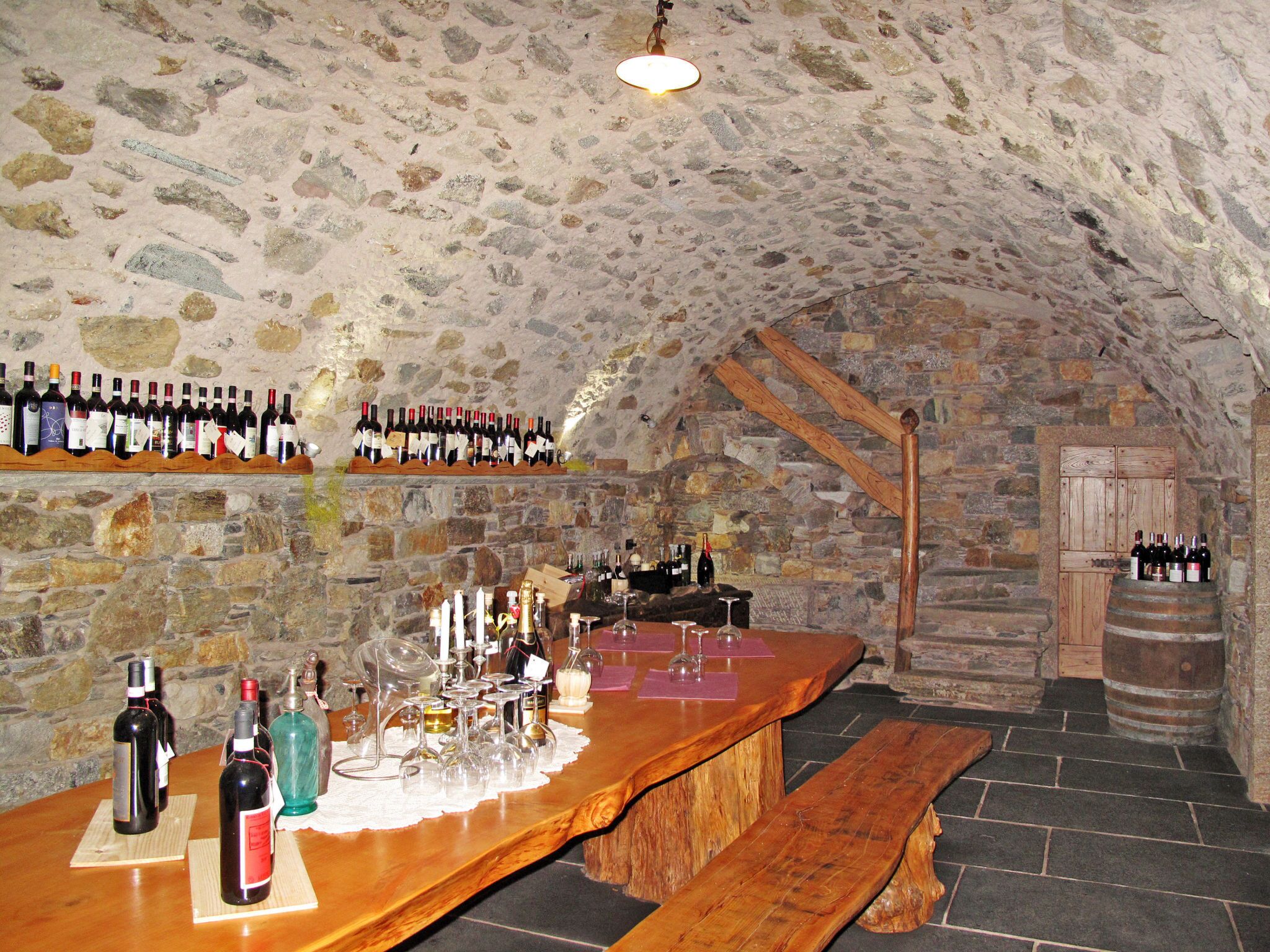 A stone-walled bar area with wooden shelves of wine bottles, a wooden bar counter, and a stone bench.