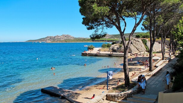 Ein Strand mit klarem, blauem Wasser, einer felsigen Küstenlinie und einigen badenden Personen.