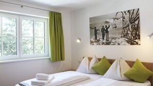 Un letto matrimoniale con lenzuola bianche e cuscini verdi, una finestra con tende verdi e un quadro appeso al muro raffigurante un paesaggio innevato.