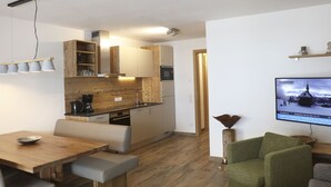 Une cuisine moderne avec des meubles en bois, un coin repas avec une table et des bancs en bois, et un salon avec un canapé vert et un téléviseur diffusant un paysage enneigé.