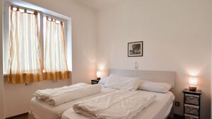 A double bed with white linens, two bedside tables with lamps, a window with curtains, and a framed picture on the wall.