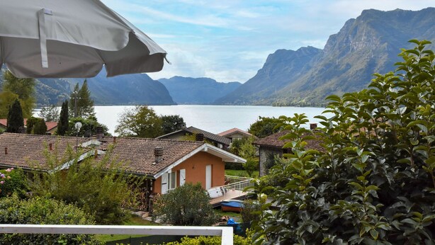 A lakeside view with mountains in the background, a house with a red roof, and a white umbrella.
