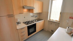 A compact kitchen with wooden cabinets, a gas stove, an oven, and a window with a curtain. - Villa Laura by Interhome (Lago d'Idro)