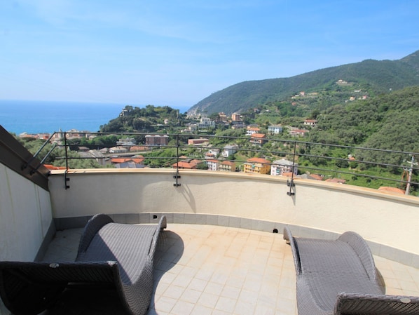 A balcony with wicker furniture overlooking a coastal town and hills.