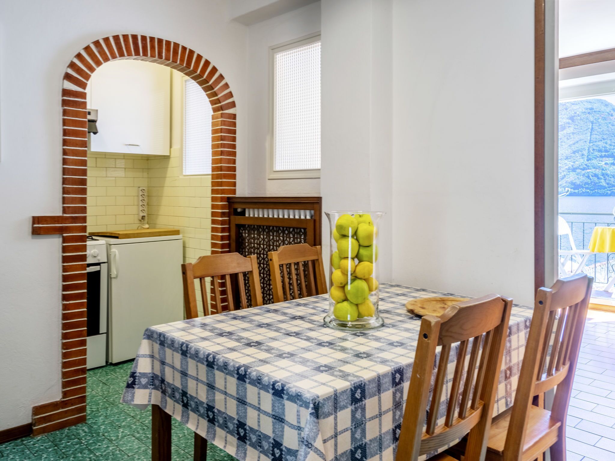 A dining area with a checkered tablecloth, a glass vase with apples, and a brick archway leading to a kitchen.