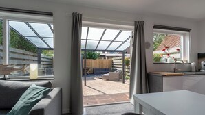 Une cuisine moderne dotée d’une grande fenêtre, un coin repas avec une table blanche et un espace salon confortable avec un canapé et un coussin vert.