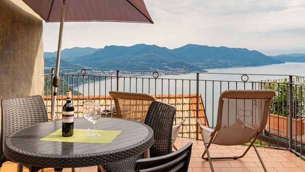 Eine Terrasse mit einem runden Tisch, zwei Stühlen und einem Sonnenschirm. Auf dem Tisch steht eine Weinflasche und zwei Gläser. Die Terrasse bietet Blick auf einen See und Berge.