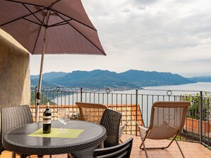 A terrace with a round table, two chairs, and an umbrella. A bottle of wine and two glasses are on the table. The terrace overlooks a lake and mountains.