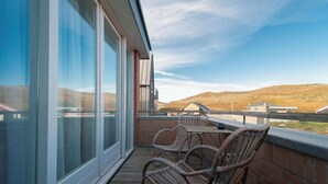 Een balkon met rieten stoelen en een tafel, grote glazen deuren en uitzicht op een heuvellandschap.