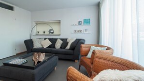 Een moderne woonkamer met een donkere bank, een glazen salontafel en rieten stoelen.