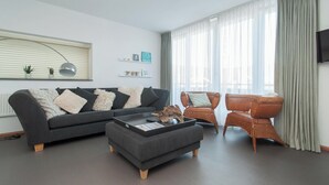 Een moderne woonkamer met een donkergrijze bank, een bijpassend voetenbankje en een rieten fauteuil. Op de bank staan twee kleine planten in potten, een aan de muur bevestigde televisie en een raam met dunne gordijnen.