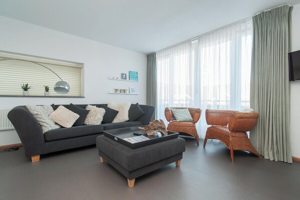 A modern living room with a dark grey sofa, a matching ottoman, and a wicker chair. There are two small potted plants on the sofa, a wall-mounted TV, and a window with sheer curtains.