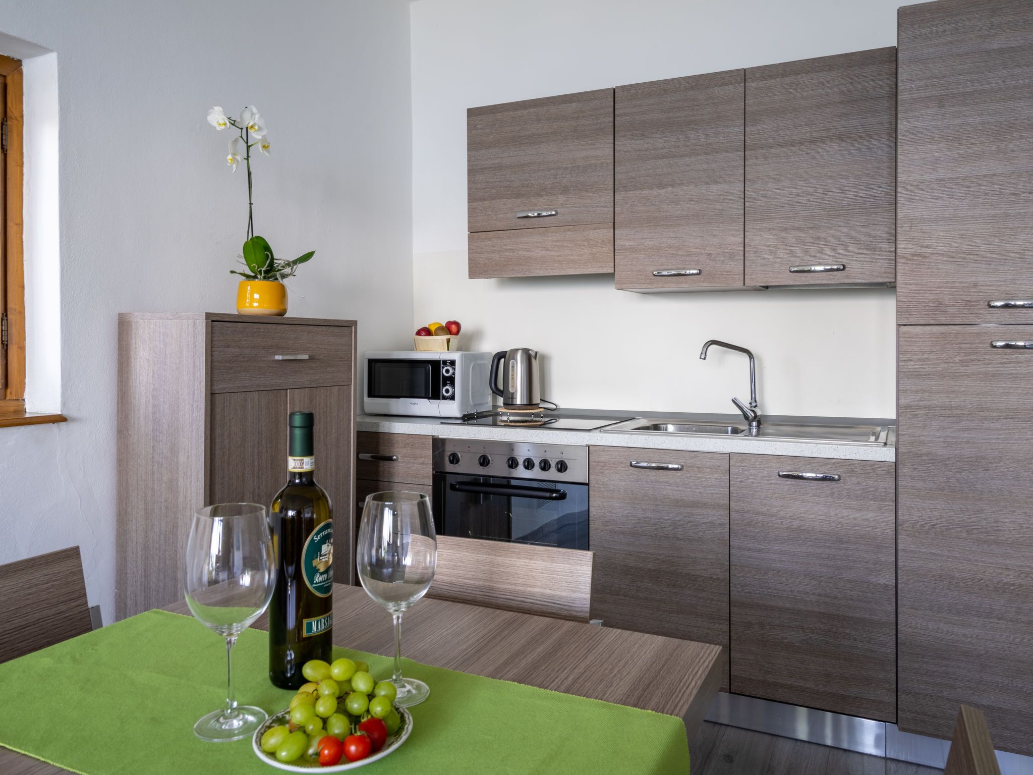 A modern kitchen with wooden cabinets, a microwave, a kettle, and a sink. There is a dining table with a green tablecloth, a bottle of wine, and a bowl of fruit.