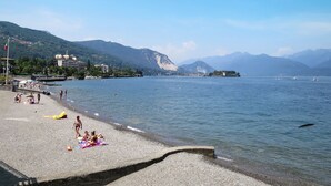 A pebbled beach with people sunbathing and playing, a clear blue sky, and mountains in the background. - Ca' delle Isole by Interhome (Stresa)
