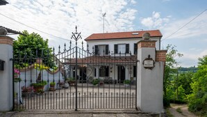 Une entrée fermée par un portail mène à une maison avec un toit en tuiles, une enseigne indiquant « Casa delle Reschie », et un jardin agrémenté de plantes et de fleurs en pot.