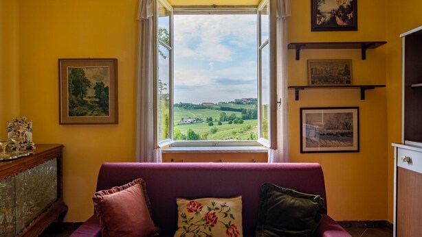 Une pièce avec un canapé violet, une fenêtre donnant sur un paysage verdoyant et des tableaux encadrés accrochés au mur.