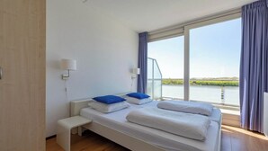 Une chambre d’hôtel moderne avec un grand lit, un banc, une fenêtre donnant sur un plan d’eau et une lampe fixée au mur.