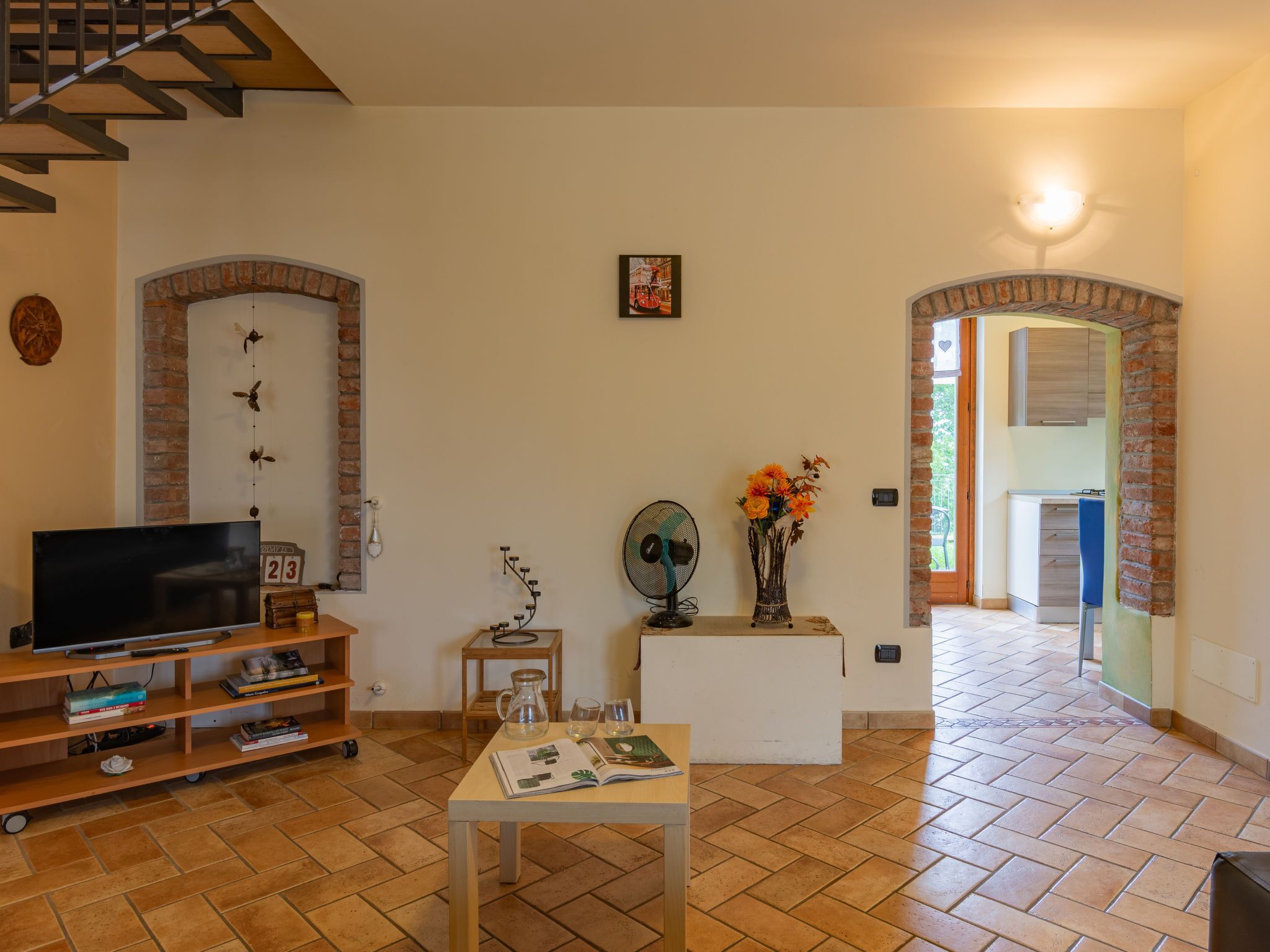 A living room with a TV, bookshelves, a coffee table, and a vase with flowers. There are brick arches and a staircase.