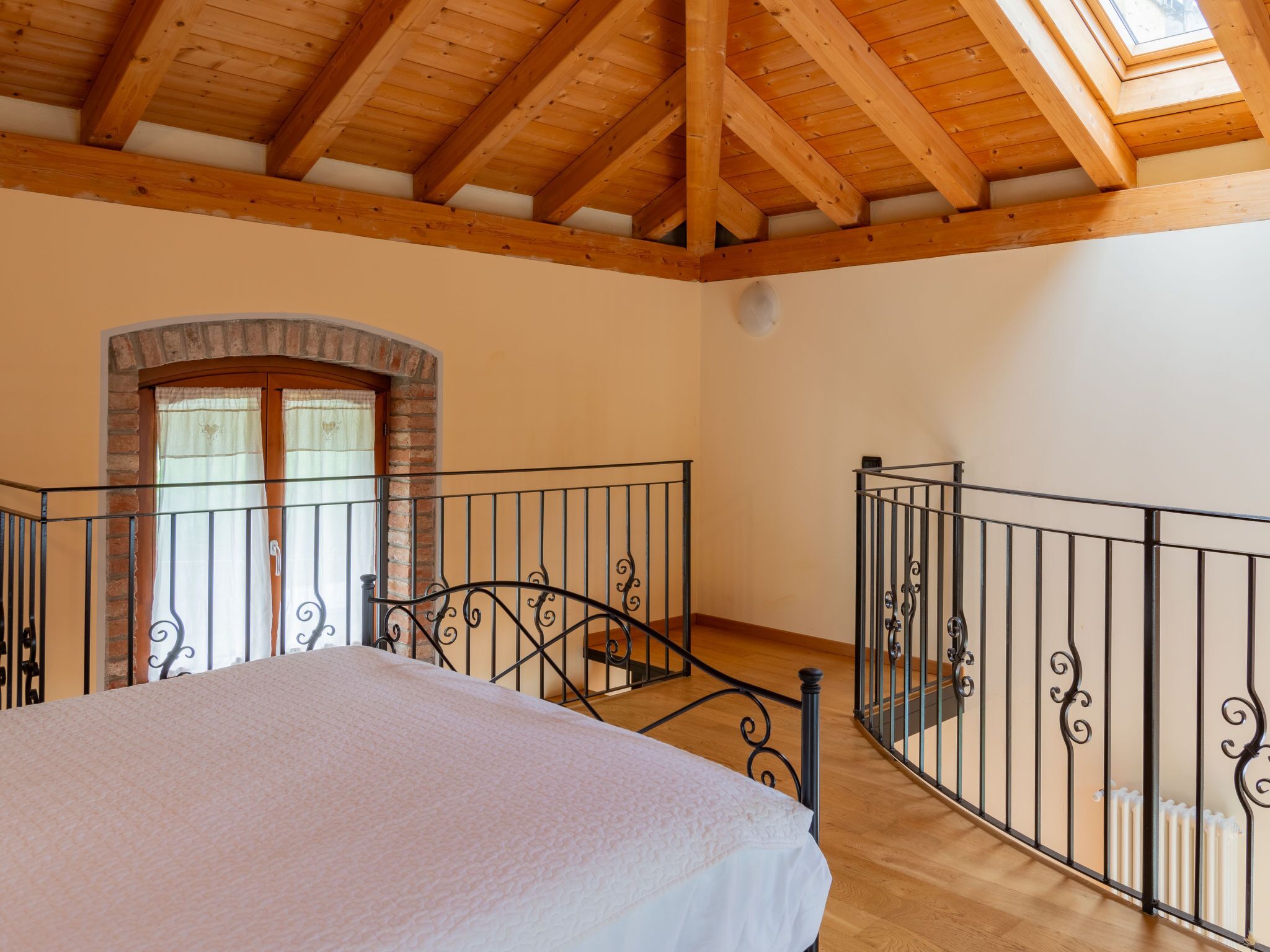 A bedroom with a wooden ceiling, a bed, a brick archway, and a balcony with metal railings.