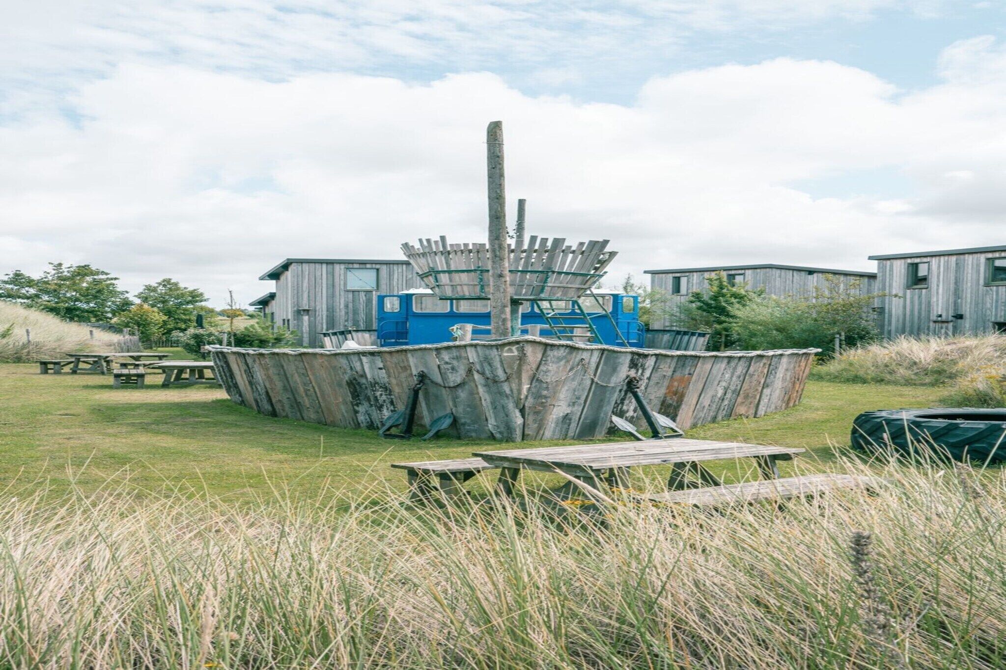 Un entorno rústico al aire libre con una gran estructura de madera que recuerda a un barco, rodeada de mesas de picnic y un campo de césped.