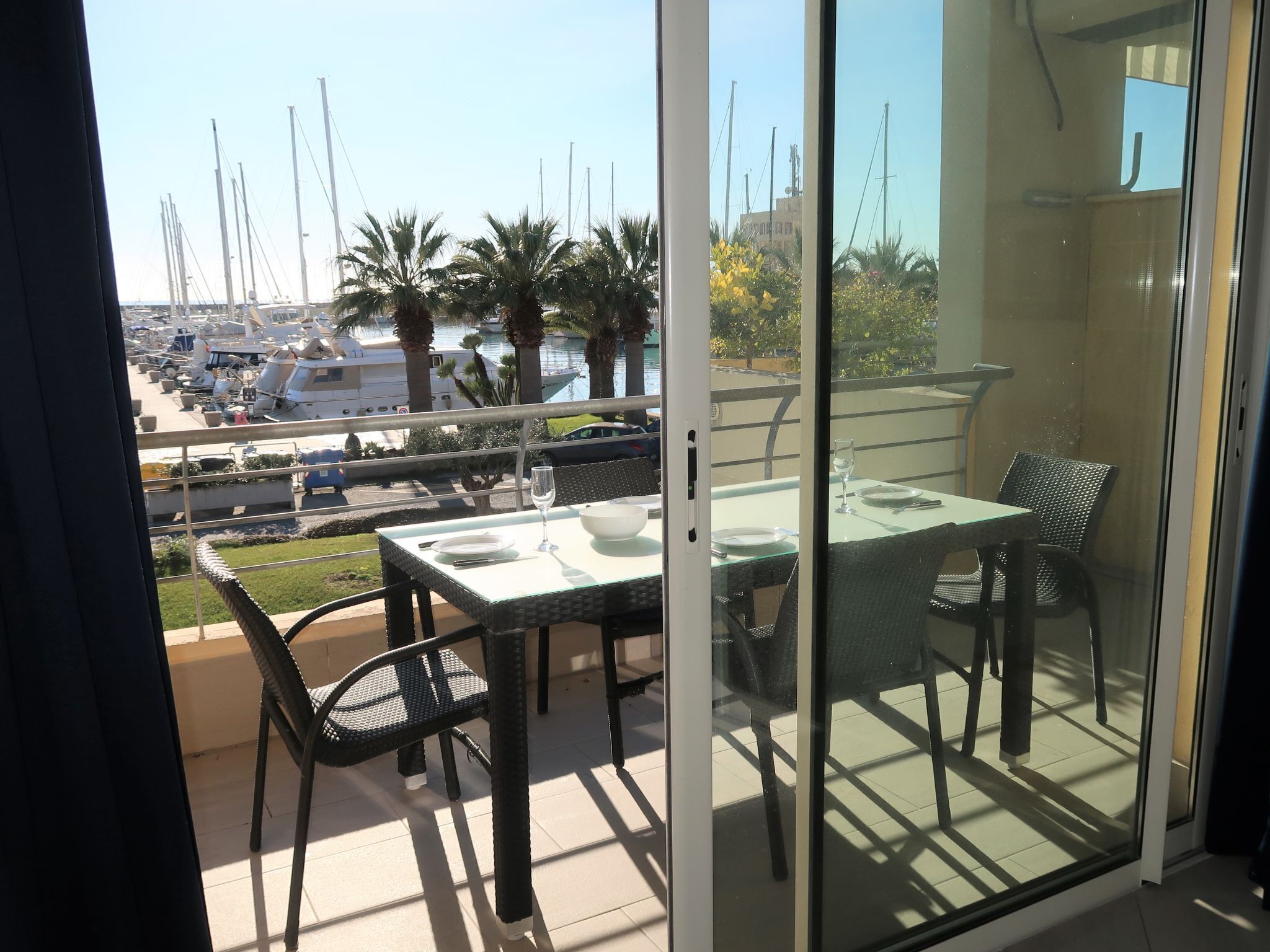 Un balcon avec une porte vitrée, une table dressée pour deux personnes et une vue sur les bateaux et les palmiers.