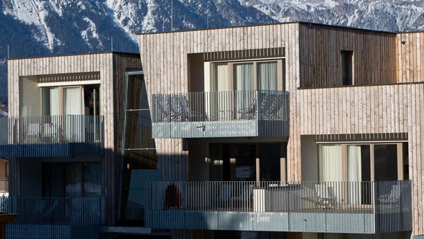 A modern wooden building with balconies and large windows, set against a backdrop of snow-capped mountains.