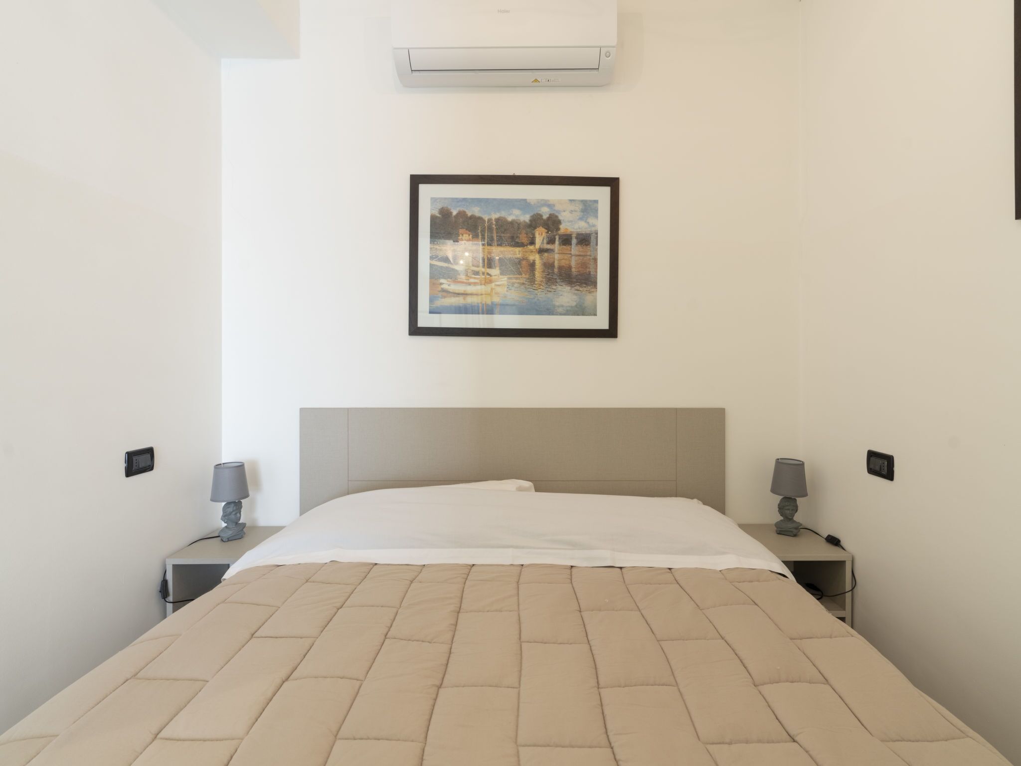 A hotel room with a bed, two bedside tables, a wall-mounted air conditioner, and a framed painting above the bed.