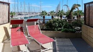 Een balkon met rode ligbedden met uitzicht op een jachthaven vol zeilboten.