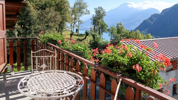 Ein Balkon mit einem Metalltisch, umgeben von Holelllaufgeländern und Kübelpflanzen mit roten Blüten.