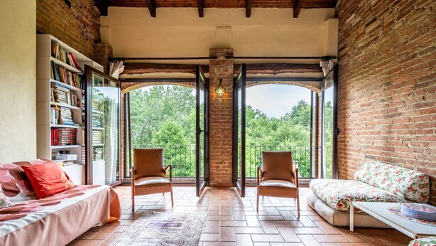 A cozy room with a brick wall, a bookshelf, a sofa, two chairs, and a large window offering a view of trees. - Vacation home Carla by Interhome (Cortazzone)
