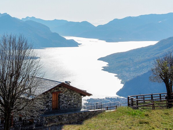 A stone building with a wooden door, a metal railing, and a view of a lake surrounded by mountains.