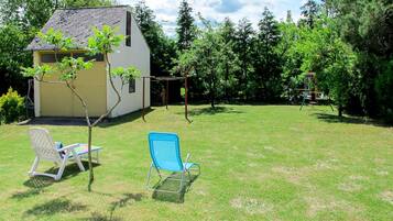 A small house with a sloped roof and a garden area with two lawn chairs and a swing set.