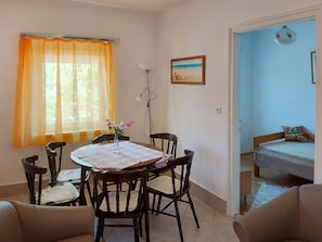 A dining area with a table, chairs, and a vase of flowers. There is a window with curtains, a lamp, and a painting on the wall.