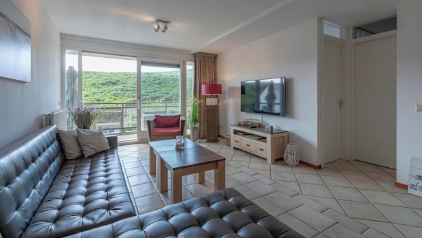 Una sala de estar moderna con un sofá de cuero, una mesa de centro de madera, un televisor de pantalla plana y un balcón con vistas a la vegetación.