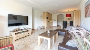 Una sala de estar moderna con un televisor de pantalla plana, una mesa de centro de madera, un sofá marrón y una zona de comedor con una cocina al fondo.
