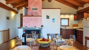 Un espace repas agrémenté d’une cheminée, de poutres apparentes en bois au plafond et d’une cuisine en arrière-plan.