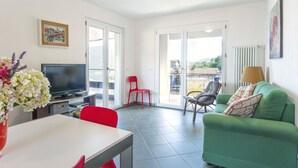 A modern living room with a green sofa, a dining table with red chairs, a TV, and a balcony with a red chair. - Madal (GRV399) by Interhome (Gravedona)