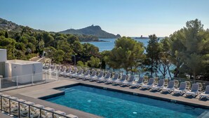 A rectangular swimming pool with clear blue water, surrounded by white lounge chairs. There is a view of a mountainous landscape and the sea in the background. - Les Chênes Verts by Interhome (Saint-Raphaël)