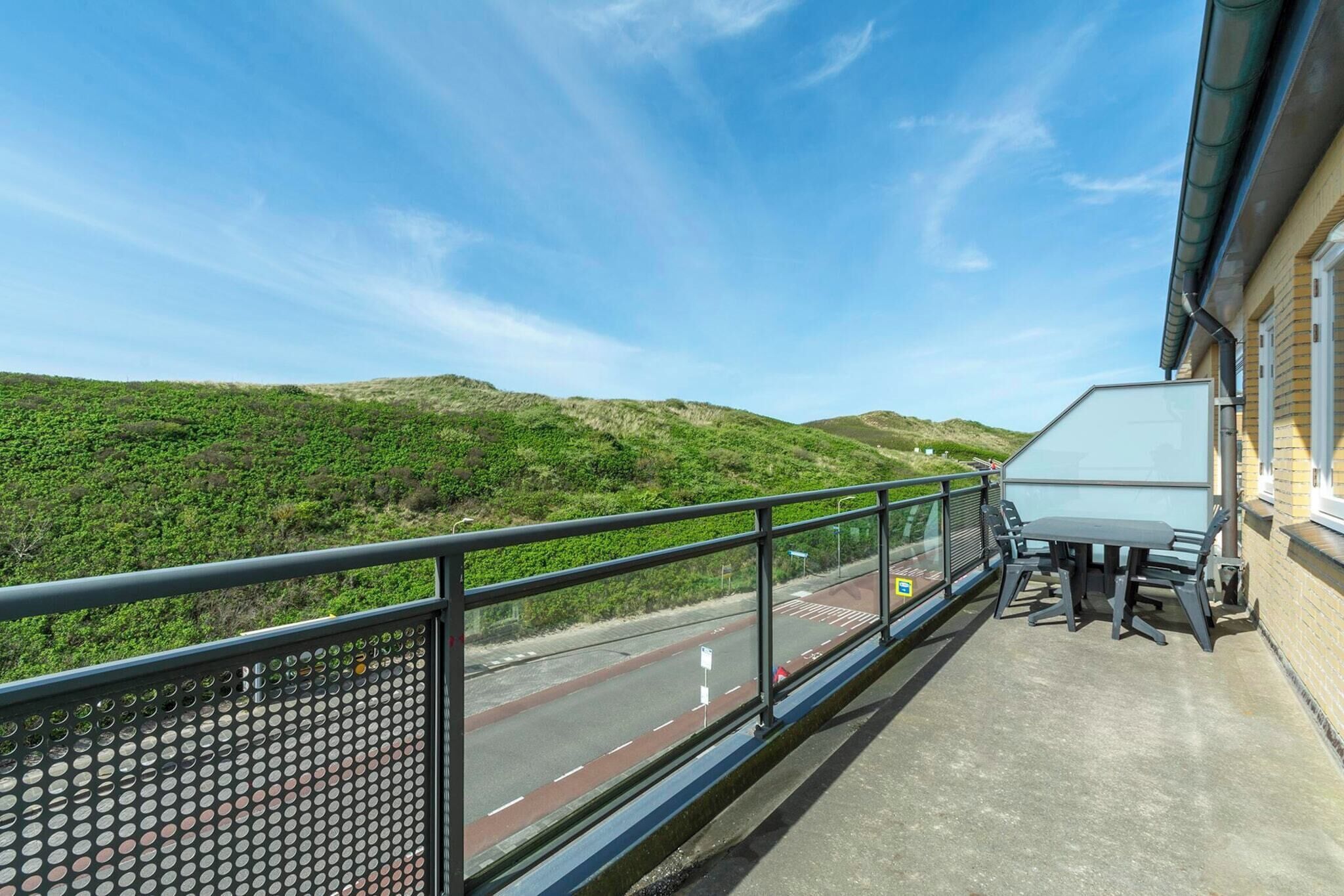 Un balcon avec une table et des chaises, donnant sur une route et des collines verdoyantes.