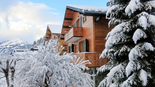 Eine Holzchalet mit Balkon, umgeben von schneebedeckten Bäumen und Bergen im Hintergrund.